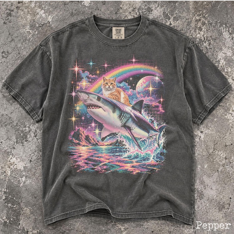May include: A charcoal gray short-sleeve t-shirt featuring a graphic print of an orange tabby cat riding a shark. The design includes a colorful rainbow, a large planet, sparkling stars, and ocean waves. The shirt has a crew neck and a label.