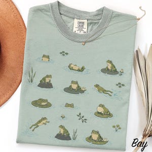 May include: A sage green t-shirt featuring a repeating graphic print of various green frogs in different poses, including swimming, sitting on lily pads, and resting on rocks, with small water lilies and reeds scattered across the fabric.
