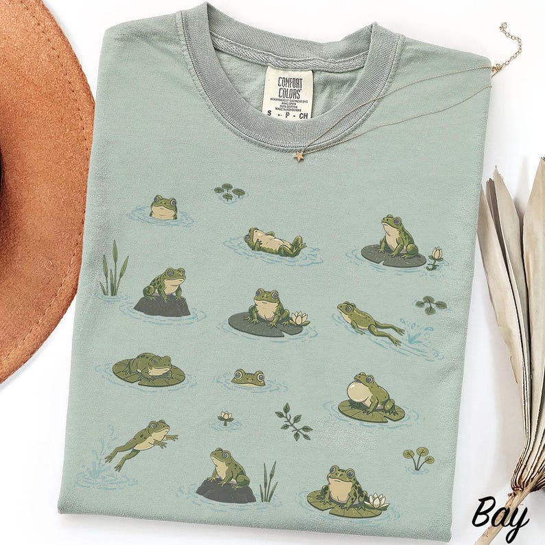 May include: A sage green t-shirt featuring a repeating graphic print of various green frogs in different poses, including swimming, sitting on lily pads, and resting on rocks, with small water lilies and reeds scattered across the fabric.