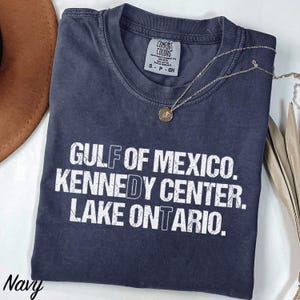 May include: A navy blue short-sleeved t-shirt with a distressed white graphic print that reads GULF OF MEXICO. KENNEDY CENTER. LAKE ONTARIO. The shirt features a crew neckline and a Comfort Colors brand label on the inside collar showing size S.