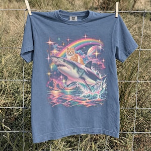 May include: A blue short-sleeve t-shirt featuring a graphic print of an orange tabby cat riding a shark. The design includes a colorful rainbow, a large planet, and sparkling stars over a sea with mountains. The shirt has a crew neckline.