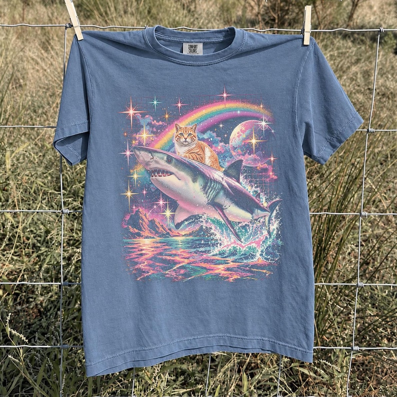May include: A blue short-sleeve t-shirt featuring a graphic print of an orange tabby cat riding a shark. The design includes a colorful rainbow, a large planet, and sparkling stars over a sea with mountains. The shirt has a crew neckline.