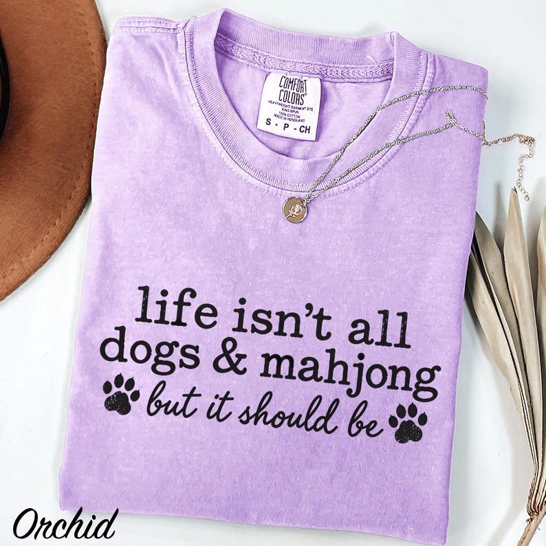 May include: A light purple short-sleeve t-shirt with a crew neckline and a Comfort Colors brand label. The shirt features black text that reads, "life isn't all dogs & mahjong but it should be," accompanied by two black paw print graphics.