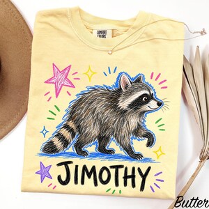 May include: A pale yellow t-shirt featuring a graphic of a raccoon with a striped tail, surrounded by colorful hand-drawn style stars and dashes. The name JIMOTHY is printed in bold black letters below the raccoon. The shirt has a Comfort Colors label.