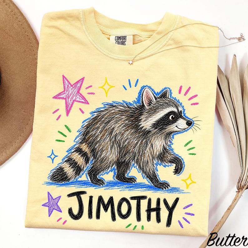 May include: A pale yellow t-shirt featuring a graphic of a raccoon with a striped tail, surrounded by colorful hand-drawn style stars and dashes. The name JIMOTHY is printed in bold black letters below the raccoon. The shirt has a Comfort Colors label.