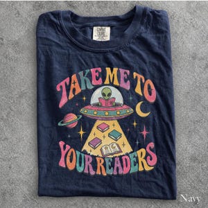 May include: A navy blue t-shirt featuring a graphic of a green alien in a flying saucer reading a book. The text reads "TAKE ME TO YOUR READERS" in colorful, retro-style lettering. The design includes planets, stars, and books floating in a beam of light.
