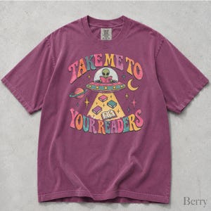 May include: A berry-colored short-sleeve t-shirt featuring a graphic print of an alien in a flying saucer reading a book. The text reads "TAKE ME TO YOUR READERS" in colorful, curved lettering, surrounded by stars, a planet, and books.
