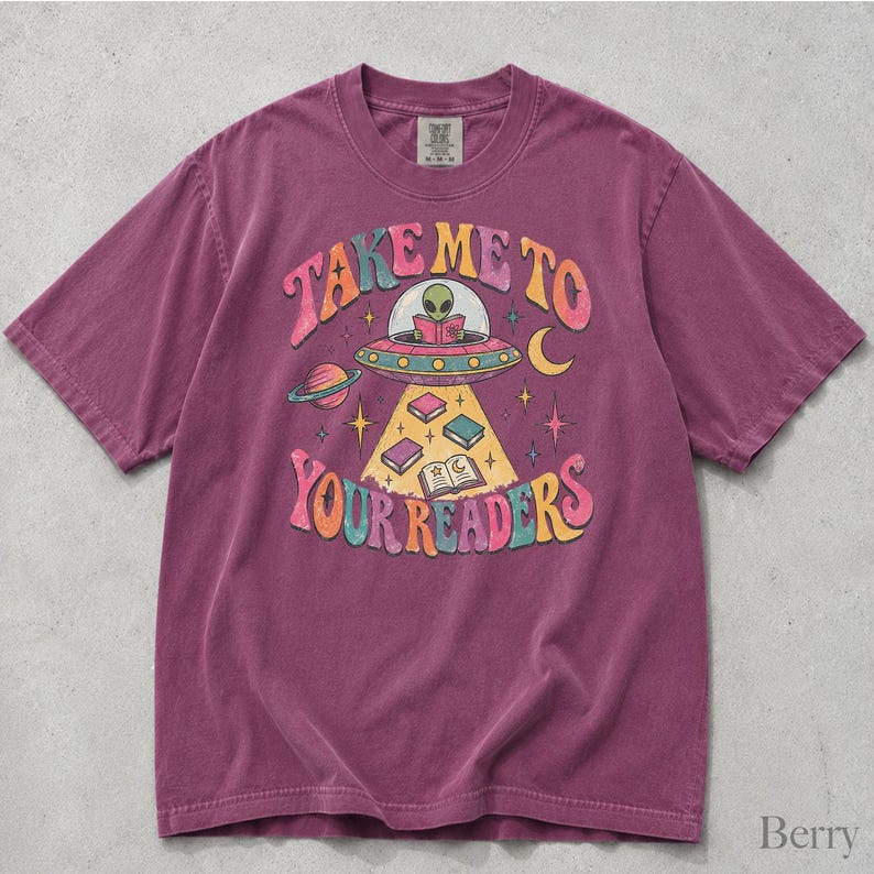 May include: A berry-colored short-sleeve t-shirt featuring a graphic print of an alien in a flying saucer reading a book. The text reads "TAKE ME TO YOUR READERS" in colorful, curved lettering, surrounded by stars, a planet, and books.