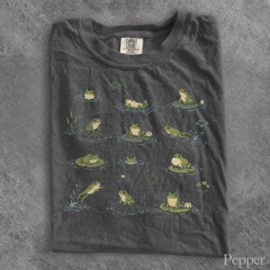 May include: A charcoal gray t-shirt featuring a graphic print of various green frogs in different poses, including sitting on lily pads, swimming, and jumping. The shirt has a crew neckline and a Comfort Colors brand label at the inner collar.