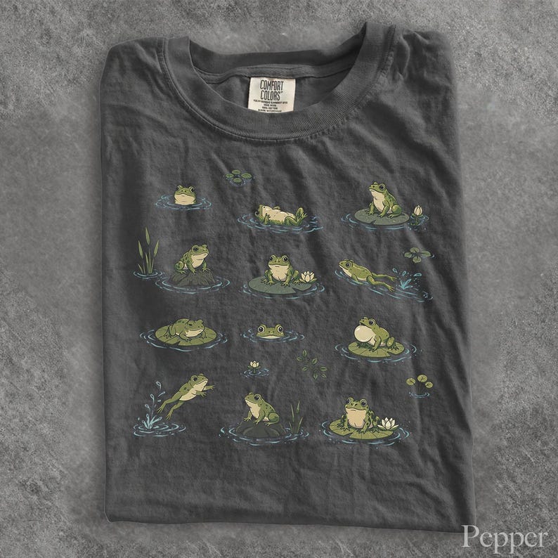May include: A charcoal gray t-shirt featuring a graphic print of various green frogs in different poses, including sitting on lily pads, swimming, and jumping. The shirt has a crew neckline and a Comfort Colors brand label at the inner collar.