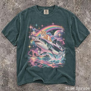 May include: A dark teal short-sleeve t-shirt featuring a graphic print of an orange tabby cat riding a shark over ocean waves. The background includes a colorful rainbow, a large moon, and sparkling stars against a dark, grid-patterned sky.