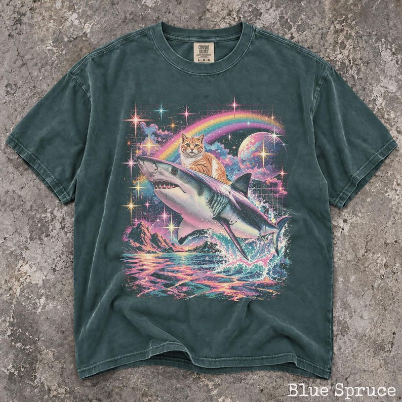 May include: A dark teal short-sleeve t-shirt featuring a graphic print of an orange tabby cat riding a shark over ocean waves. The background includes a colorful rainbow, a large moon, and sparkling stars against a dark, grid-patterned sky.