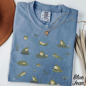 May include: A blue pigment-dyed t-shirt featuring a repeating pattern of green frogs in various poses on lily pads and in water. The inner neck label reads Comfort Colors, Heavyweight Garment Dye, 100% Cotton, Made in Honduras, size S-P-CH.
