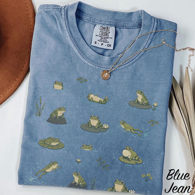 May include: A blue pigment-dyed t-shirt featuring a repeating pattern of green frogs in various poses on lily pads and in water. The inner neck label reads Comfort Colors, Heavyweight Garment Dye, 100% Cotton, Made in Honduras, size S-P-CH.