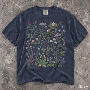 May include: A navy blue short-sleeve t-shirt featuring a detailed botanical print of various wildflowers and plants in shades of pink, purple, yellow, and white. The shirt has a crew neckline and a faded, washed-out fabric texture throughout.