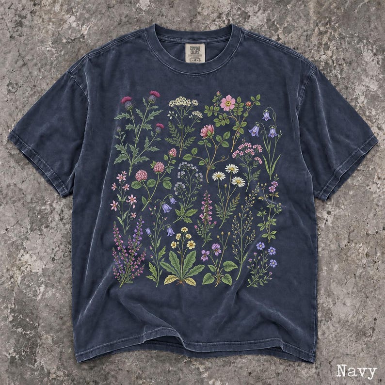 May include: A navy blue short-sleeve t-shirt featuring a detailed botanical print of various wildflowers and plants in shades of pink, purple, yellow, and white. The shirt has a crew neckline and a faded, washed-out fabric texture throughout.