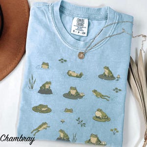 May include: A light blue t-shirt featuring a repeating pattern of green frogs in various poses, including swimming, sitting on lily pads, and floating. The shirt has a crew neckline with a Comfort Colors label and a silver-toned pendant necklace.