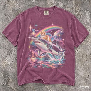 May include: A berry-colored short-sleeve t-shirt featuring a graphic print of an orange tabby cat riding a shark through a cosmic scene. The design includes a rainbow, a planet, and sparkling stars above a mountain range and ocean waves.