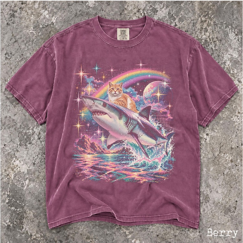 May include: A berry-colored short-sleeve t-shirt featuring a graphic print of an orange tabby cat riding a shark through a cosmic scene. The design includes a rainbow, a planet, and sparkling stars above a mountain range and ocean waves.