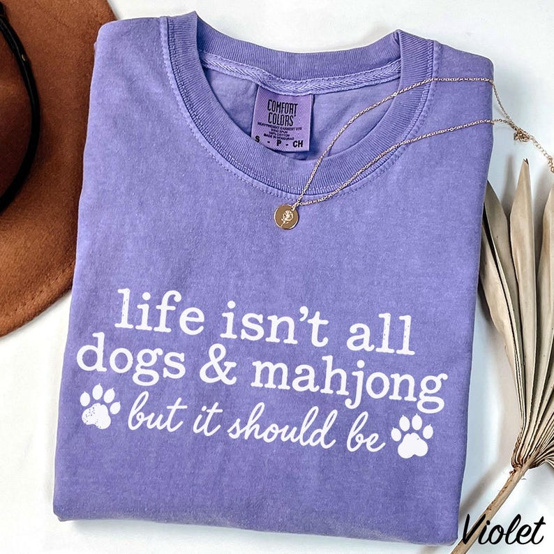 May include: A violet short-sleeved t-shirt with a crew neckline and a Comfort Colors label. The front features white text that reads, "life isn't all dogs & mahjong but it should be," accompanied by two white paw print graphics on the sides.