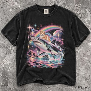May include: A black short-sleeve t-shirt featuring a graphic print of an orange tabby cat riding a shark. The design includes a colorful rainbow, a moon, stars, and ocean waves in vibrant hues. The shirt has a crew neckline and a Comfort Colors label.