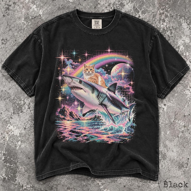 May include: A black short-sleeve t-shirt featuring a graphic print of an orange tabby cat riding a shark. The design includes a colorful rainbow, a moon, stars, and ocean waves in vibrant hues. The shirt has a crew neckline and a Comfort Colors label.