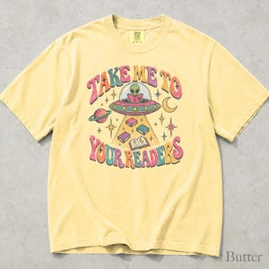 May include: A pale yellow short-sleeve t-shirt featuring a graphic of an alien in a flying saucer reading a book. The text "TAKE ME TO YOUR READERS" is printed in a curved, colorful font. The design includes planets, stars, and books.