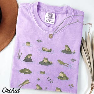 May include: A light purple short-sleeve t-shirt featuring a repeating pattern of green frogs in various poses on lily pads and in water. The inner neck label reads Comfort Colors, Heavyweight Garment Dye, Ring Spun, 100% Cotton, Made in Honduras, S-P-CH.