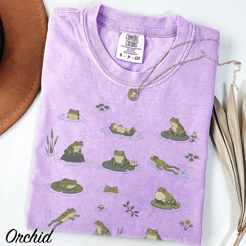 May include: A light purple short-sleeve t-shirt featuring a repeating pattern of green frogs in various poses on lily pads and in water. The inner neck label reads Comfort Colors, Heavyweight Garment Dye, Ring Spun, 100% Cotton, Made in Honduras, S-P-CH.