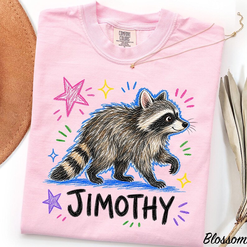 May include: A light pink t-shirt featuring a graphic of a raccoon with a striped tail. The design includes colorful hand-drawn style stars and dashes in pink, yellow, blue, and green. The name JIMOTHY is printed in large black letters below.