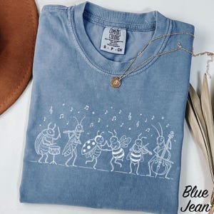 Op de afbeelding: Een blauw T-shirt met korte mouwen voorzien van een witte lijntekening van een groep insecten die muziekinstrumenten bespelen, waaronder een kever op drums, een sprinkhaan op fluit, een lieveheersbeestje, een bij en een krekel op cello.