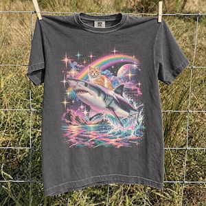 May include: A charcoal gray short-sleeve t-shirt featuring a graphic print of an orange tabby cat riding on the back of a great white shark. The design includes a colorful rainbow, a large planet, sparkling stars, and ocean waves in vibrant hues.