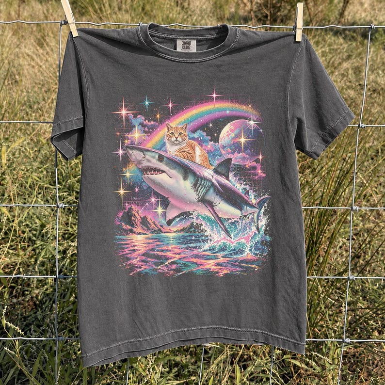 May include: A charcoal gray short-sleeve t-shirt featuring a graphic print of an orange tabby cat riding on the back of a great white shark. The design includes a colorful rainbow, a large planet, sparkling stars, and ocean waves in vibrant hues.