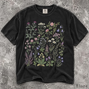 May include: A black short-sleeve t-shirt featuring a botanical print of various wildflowers in shades of pink, purple, yellow, and white. The shirt has a crew neckline and a label at the inner collar that reads Comfort Colors with size L-G-G.