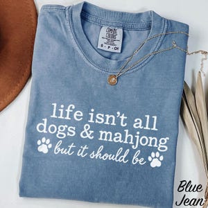 May include: A blue short-sleeve t-shirt with a white graphic print that reads, "life isn't all dogs & mahjong but it should be" with two paw prints. The shirt features a Comfort Colors brand label at the neck and a small gold-toned pendant necklace.