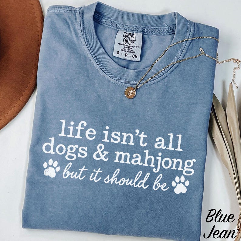 May include: A blue short-sleeve t-shirt with a white graphic print that reads, "life isn't all dogs & mahjong but it should be" with two paw prints. The shirt features a Comfort Colors brand label at the neck and a small gold-toned pendant necklace.