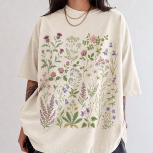 May include: An oversized cream-colored t-shirt featuring a detailed botanical print on the front. The graphic displays a variety of colorful wildflowers, including purple thistles, pink roses, blue bellflowers, and white daisies, arranged in a grid.