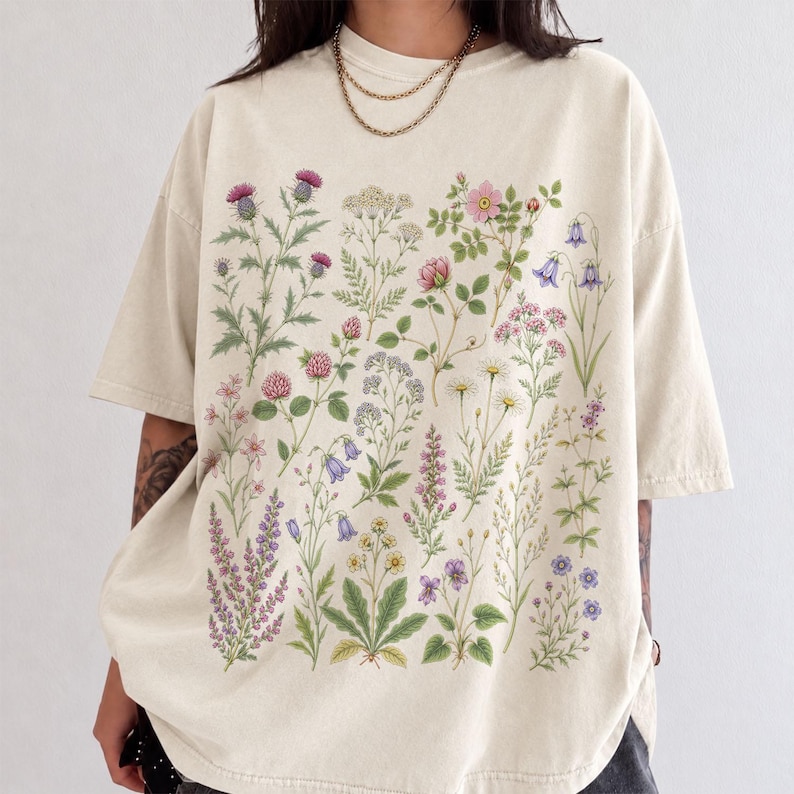 May include: An oversized cream-colored t-shirt featuring a detailed botanical print on the front. The graphic displays a variety of colorful wildflowers, including purple thistles, pink roses, blue bellflowers, and white daisies, arranged in a grid.