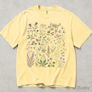 May include: A pale yellow short-sleeve t-shirt featuring a detailed botanical print on the front. The graphic displays an array of colorful wildflowers, including purple thistles, pink roses, blue bellflowers, and white daisies on green stems.