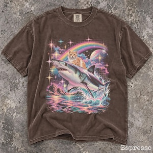May include: A brown short-sleeve t-shirt featuring a graphic print of an orange tabby cat riding a shark over ocean waves. The design includes a rainbow, a planet, and sparkling stars in the background. The shirt has a crew neckline and a label.