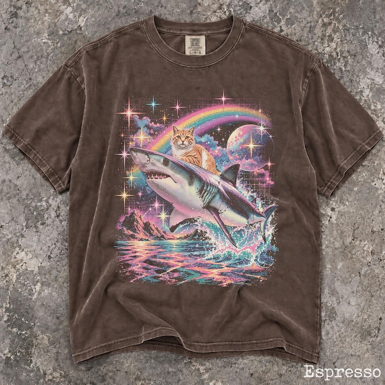 May include: A brown short-sleeve t-shirt featuring a graphic print of an orange tabby cat riding a shark over ocean waves. The design includes a rainbow, a planet, and sparkling stars in the background. The shirt has a crew neckline and a label.