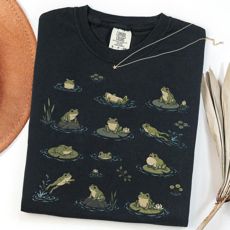 May include: A black t-shirt featuring a graphic print of various green frogs in different poses, including sitting on lily pads, swimming, and resting on rocks. The shirt has a crew neckline and a Comfort Colors brand label at the inner collar.