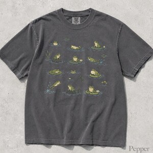 May include: A charcoal gray short-sleeve t-shirt featuring a graphic print of various green frogs in different poses, some sitting on lily pads, with small water ripples and aquatic plants scattered across the front of the garment.