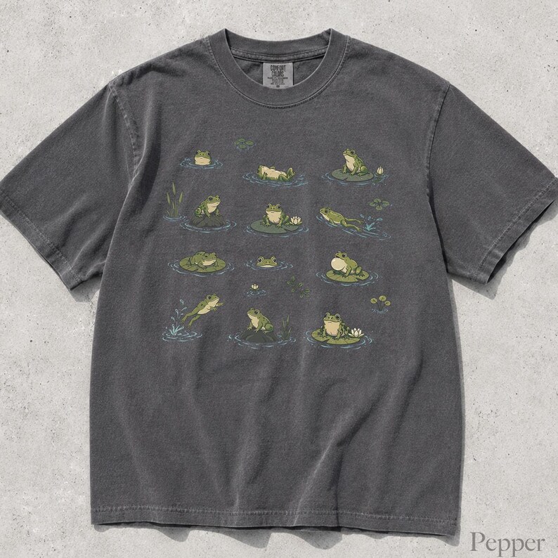 May include: A charcoal gray short-sleeve t-shirt featuring a graphic print of various green frogs in different poses, some sitting on lily pads, with small water ripples and aquatic plants scattered across the front of the garment.