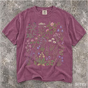 May include: A berry-colored short-sleeve t-shirt featuring a detailed botanical print of various wildflowers and plants in shades of green, pink, blue, white, and yellow. The shirt has a crew neckline and a label at the inner neck area.