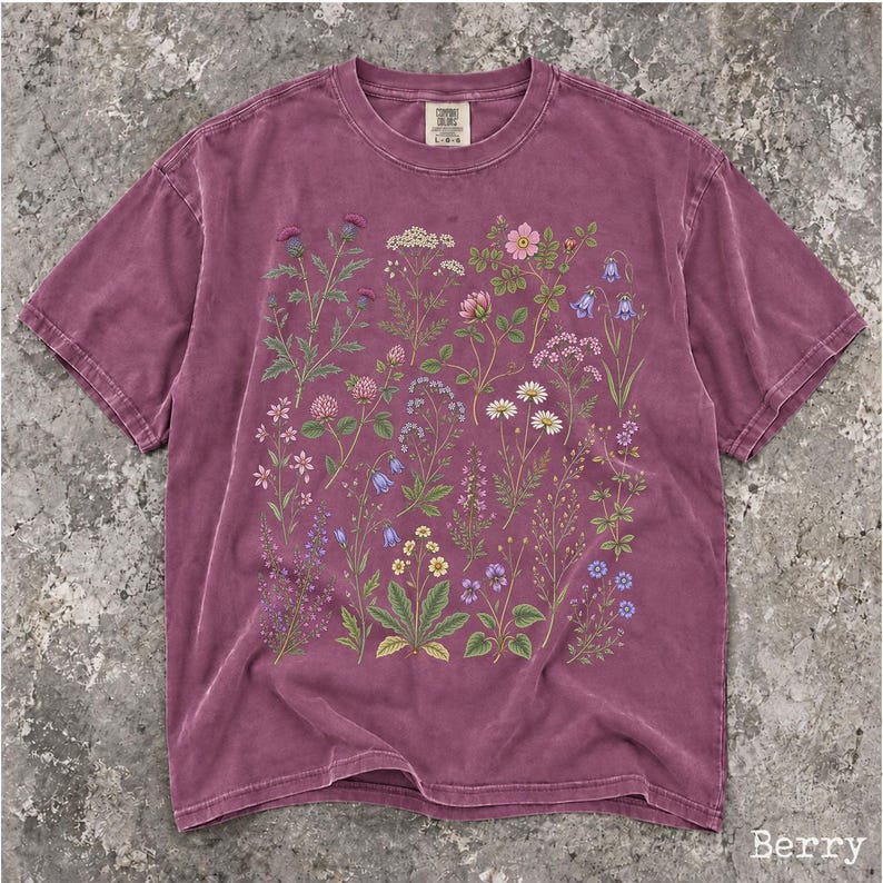 May include: A berry-colored short-sleeve t-shirt featuring a detailed botanical print of various wildflowers and plants in shades of green, pink, blue, white, and yellow. The shirt has a crew neckline and a label at the inner neck area.