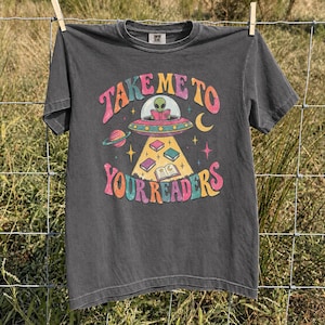 May include: A charcoal gray short-sleeve t-shirt featuring a colorful graphic of an alien in a flying saucer reading a book. The text reads "TAKE ME TO YOUR READERS" in a retro font, surrounded by planets, stars, and floating books.