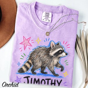 May include: A light purple short-sleeve t-shirt featuring a graphic illustration of a raccoon. The design includes colorful hand-drawn style stars and the name JIMOTHY printed in black below the raccoon. The shirt has a Comfort Colors brand label.