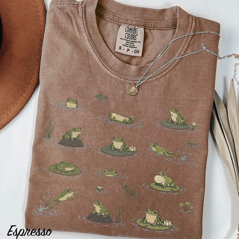 May include: A brown short-sleeve t-shirt featuring a graphic print of various green frogs in different poses on lily pads and in water. The shirt has a Comfort Colors brand label at the inner neckline and a silver-toned necklace with a round pendant.
