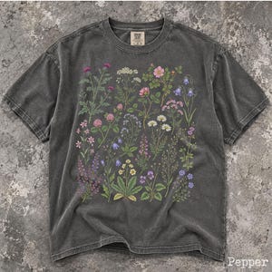 May include: A charcoal gray short-sleeve t-shirt featuring a detailed botanical print of various wildflowers in shades of pink, purple, yellow, and white. The shirt has a crew neckline and a Comfort Colors brand label printed on the inner collar.